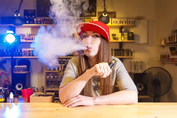 Jeune jolie femme en bonnet rouge fume une cigarette électronique à la boutique de vape — Photo