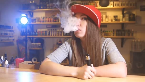 Una joven guapa con gorra roja fuma un cigarrillo electrónico en la tienda de vapores. Primer plano. . — Vídeos de Stock