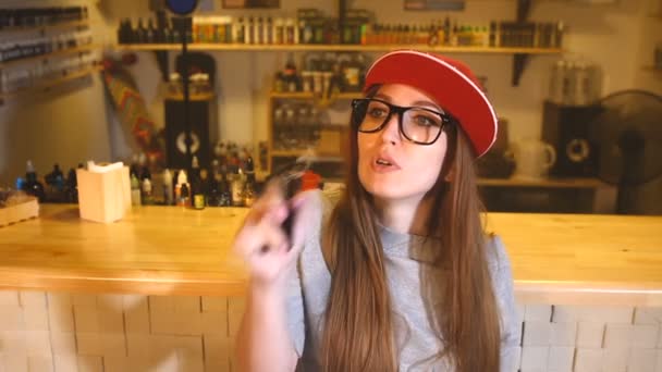 Una joven guapa con gorra roja fuma un cigarrillo electrónico en la tienda de vapores. Primer plano. . — Vídeos de Stock