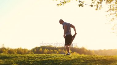 parkta egzersiz için hazırlanıyor germe egzersiz yaparak genç atletik atlet. gün batımı, tonda video