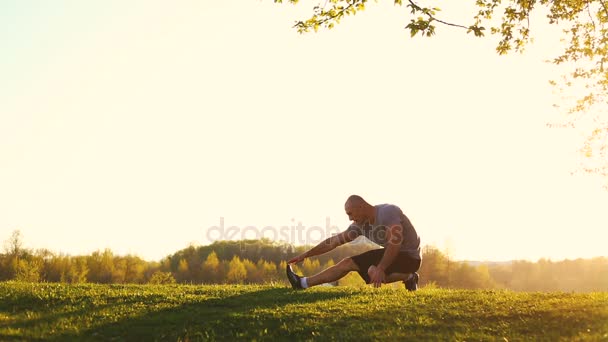 Jonge sportieve loper doen stretching oefening, voorbereiding op training in het park. zonsondergang, getinte video — Stockvideo
