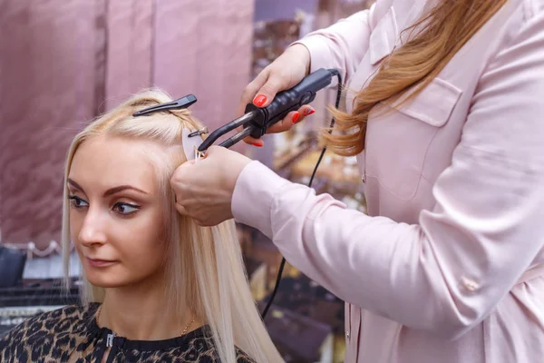 Hair extensions procedure. Hairdresser does hair extensions to young girl, blonde in a beauty salon. Selective focus.