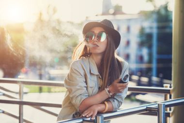 Genç ve güzel hipster kadın siyah şapka vape ecig, gün batımında vaping aygıt. Tonlu görüntü.