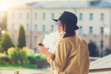 Genç ve güzel hipster kadın siyah şapka vape ecig, gün batımında vaping aygıt. Tonlu görüntü.