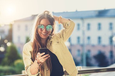 Şık genç kadın, bir telefon holding eller güzel. Gün batımı arka plan, güneşli bir gün, iyi bir hava, güneş gözlüğü, serin aksesuarları.