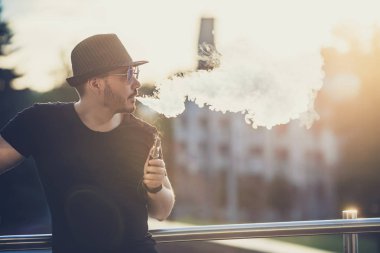 Arka planda, akşam gün batımını şehrin üzerinde bir sakal eğlenceli Vaporizers, Şapkalı modern mutlu genç adam. tonlu görüntü.
