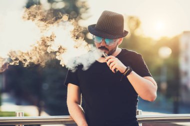 Arka planda, akşam gün batımını şehrin üzerinde bir sakal eğlenceli Vaporizers, Şapkalı modern mutlu genç adam. tonlu görüntü.