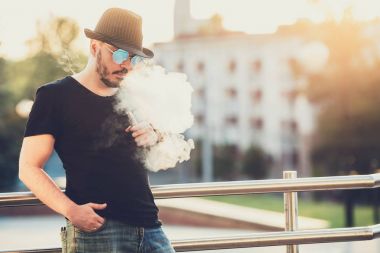 Arka planda, akşam gün batımını şehrin üzerinde bir sakal eğlenceli Vaporizers, Şapkalı modern mutlu genç adam. tonlu görüntü.