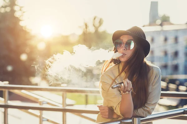 Hübsche junge Hipsterfrau mit schwarzem Hut, die im Sonnenuntergang dampft. Getöntes Image. — Stockfoto