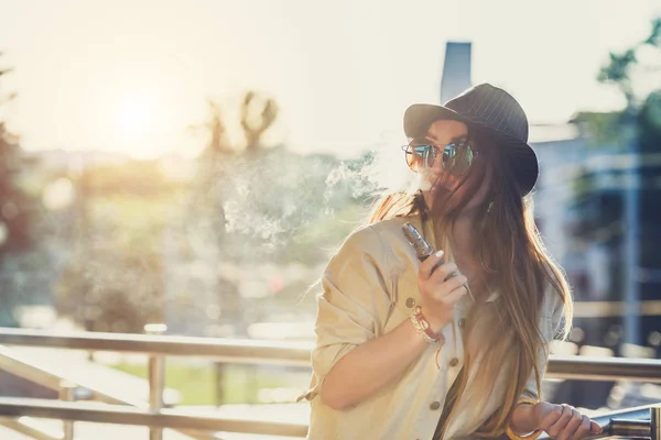 Hübsche junge Hipsterfrau mit schwarzem Hut, die im Sonnenuntergang dampft. Getöntes Image. — Stockfoto