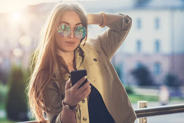 Young stylish pretty Woman, hands holding a phone. Sunset background, Sunny day,good weather, sunglasses, cool accessories. — Stock Photo, Image