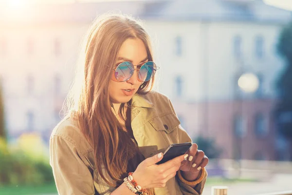 Young stylish pretty Woman, hands holding a phone. Sunset background, Sunny day,good weather, sunglasses, cool accessories. — Stock Photo, Image