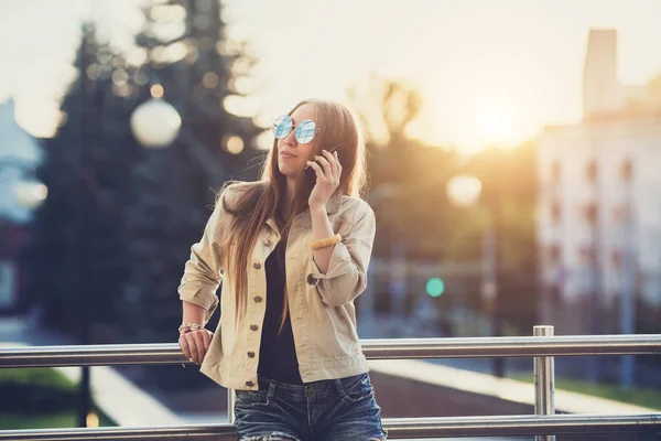 Junge stylische hübsche Frau, die Hände halten ein Telefon. Sonnenuntergang, sonniger Tag, gutes Wetter, Sonnenbrille, coole Accessoires. — Stockfoto