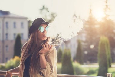 Genç ve güzel hipster kadın siyah şapka vape ecig, gün batımında vaping aygıt. Tonlu görüntü.