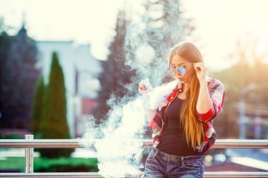Genç güzel hipster kadın vaping ve Buhar bulutu serbest bırakır. Akşam güneşin şehri. Tonlu görüntü.