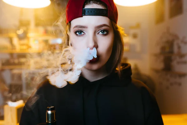 Young pretty woman in red cap smoke an electronic cigarette at the vape shop. Hip-hop style. Closeup. — Stock Photo, Image