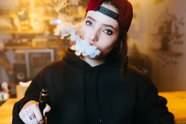 Fiatal szép nő a piros sapkát füst egy elektronikus cigaretta a vape boltban. Hip-hop stílusban. Vértes. — Stock Fotó