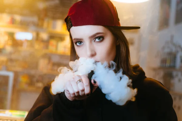 Une jeune jolie femme en bonnet rouge fume une cigarette électronique à la boutique de singes. Style hip-hop. Gros plan . — Photo