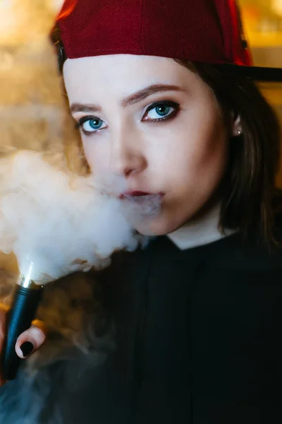 Uma jovem bonita de boné vermelho fuma um cigarro eletrônico na loja de vape. Estilo hip-hop. Fechar. . — Fotografia de Stock