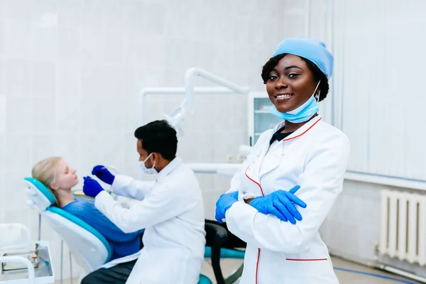 Joven dentista afroamericana sonriente delante del dentista revisando los dientes a la paciente en la clínica. Concepto de clínica dental . — Foto de Stock