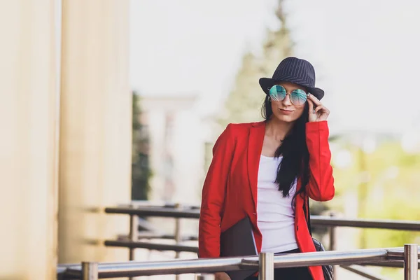 Портрет счастливой хипстерши молодой женщины с ноутбуком в солнечных очках, черной шляпе и красной куртке. Студентка с ноутбуком в кампусе университета. Тонированное изображение . — стоковое фото