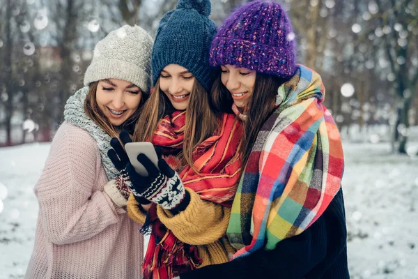 Three women friends outdoors in knitted hats using mobile phone on a snowy cold weather. Group of young female friends outdoors in winter park.