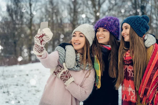 在寒冷的雪天里，三个戴着针织帽子的女性朋友在户外用手机聊天。 一群年轻的女性朋友在冬季公园里享受户外自拍. — 图库照片