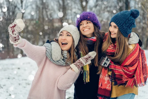 Три подруги на улице в вязаных шляпах используют мобильный телефон в снежную холодную погоду. Группа молодых подруг, наслаждающихся селфи на природе в зимнем парке . — стоковое фото