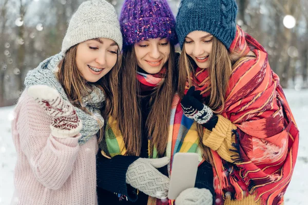 Three women friends outdoors in knitted hats using mobile phone on a snowy cold weather. Group of young female friends outdoors in winter park.