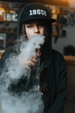 Şapkalı genç bir kadın vape dükkanında elektronik sigara içiyor. Hip-hop tarzı. Yakın çekim..