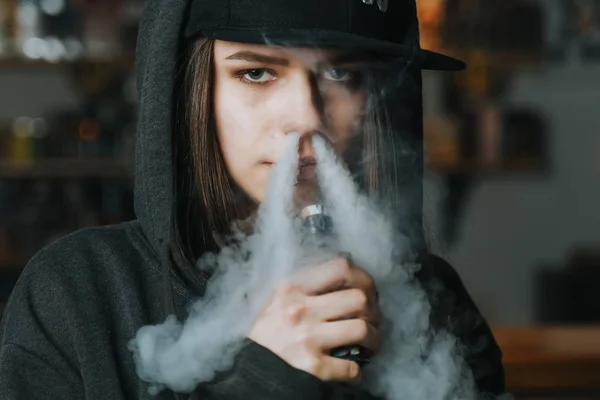 Jovem bonita de boné fuma um cigarro eletrônico na loja de vape. Estilo hip-hop. Fechar. . — Fotografia de Stock