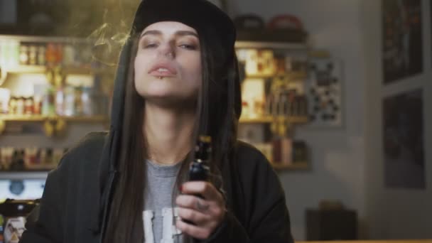 Ung söt kvinna i mössa röker en elektronisk cigarett i ångbutiken. Hip-hop-stil. Närbild. — Stockvideo