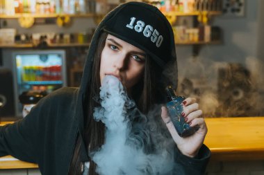 Şapkalı genç bir kadın vape dükkanında elektronik sigara içiyor. Hip-hop tarzı. Yakın çekim..