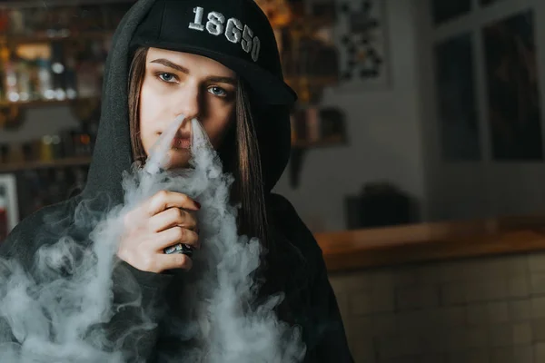 Jovem bonita de boné fuma um cigarro eletrônico na loja de vape. Estilo hip-hop. Fechar. . — Fotografia de Stock
