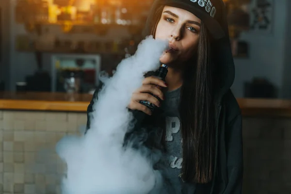 Jovem bonita de boné fuma um cigarro eletrônico na loja de vape. Estilo hip-hop. Fechar. . — Fotografia de Stock