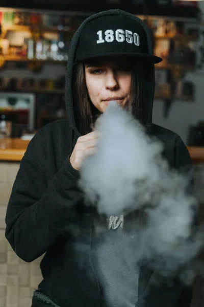Молода красива жінка в кепці курить електронну сигарету у вейп-шопі. Стиль хіп-хоп. крупним планом . — стокове фото