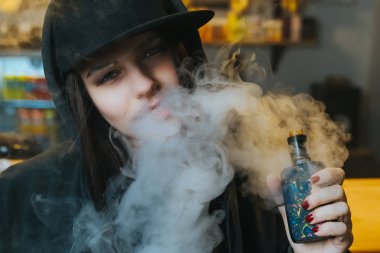Şapkalı genç bir kadın vape dükkanında elektronik sigara içiyor. Hip-hop tarzı. Yakın çekim..