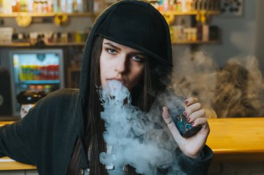 Şapkalı genç bir kadın vape dükkanında elektronik sigara içiyor. Hip-hop tarzı. Yakın çekim..