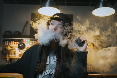 Şapkalı genç bir kadın vape dükkanında elektronik sigara içiyor. Hip-hop tarzı. Yakın çekim..