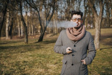 Güneş gözlüklü, kendinden emin genç sakallı erkek elektronik sigara içiyor. Genç, yakışıklı, sakallı bir hippi parkta elektronik sigara içiyor. Kapat..