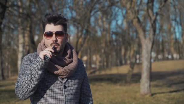 Ein junger gutaussehender, bärtiger Hipster-Mann, der im Frühjahr im Park eine elektronische Zigarette verdampft. Nahaufnahme. — Stockvideo