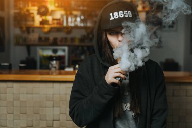 Şapkalı genç bir kadın vape dükkanında elektronik sigara içiyor. Hip-hop tarzı. Yakın çekim..
