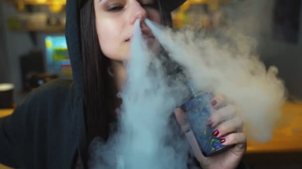 Una joven guapa con gorra fuma un cigarrillo electrónico en la tienda de vapores. Estilo hip-hop. Primer plano. . — Vídeos de Stock