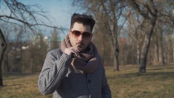 Un jeune hipster barbu vaporisant une cigarette électronique dans le parc au printemps. Gros plan . — Video