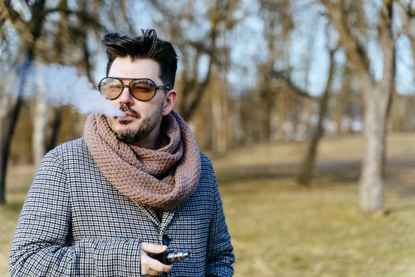 Confident young bearded male in sunglasses vape electronic cigarette. A young handsome bearded hipster man vaping e-cigarette outdoors in the park. Close up.