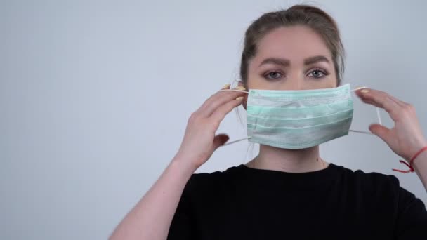 Kvinna bär mask och ser till kamera på vit bakgrund. Ett pandemiskt, beskärt porträtt av europeiska unga kvinnor som bär skyddsmask. Coronavirus-konceptet. Hälsovård och sjukvård. — Stockvideo