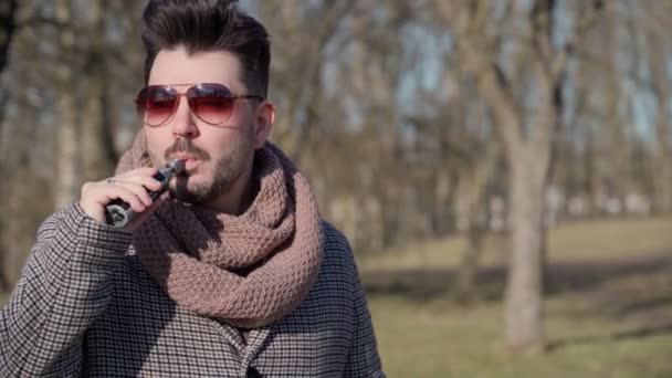 Mladý pohledný vousatý hipster, který na jaře pije v parku elektronickou cigaretu. Zavřít. — Stock video