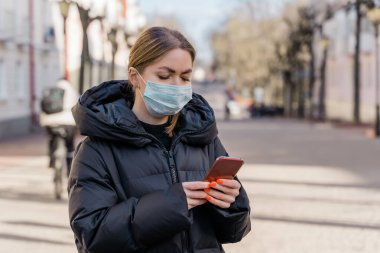 Avrupa sokaklarında akıllı telefon kullanan koruyucu tıbbi maskeli bir kadının portresi. Sağlık ve güvenlik, N1H1 koronavirüs, covid-19, virüs koruma ve tıbbi konsept. Sol tarafı çevir.