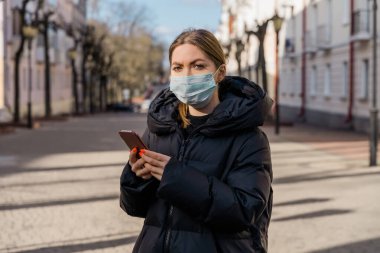 Avrupa sokaklarında akıllı telefon kullanan koruyucu tıbbi maskeli bir kadının portresi. Sağlık ve güvenlik, N1H1 koronavirüs, covid-19, virüs koruma ve tıbbi konsept. Sol tarafı çevir.
