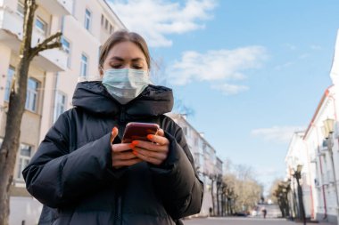 Avrupa sokaklarında akıllı telefon kullanan koruyucu tıbbi maskeli bir kadının portresi. Sağlık ve güvenlik, N1H1 koronavirüs, covid-19, virüs koruma ve tıbbi konsept. Sol tarafı çevir.
